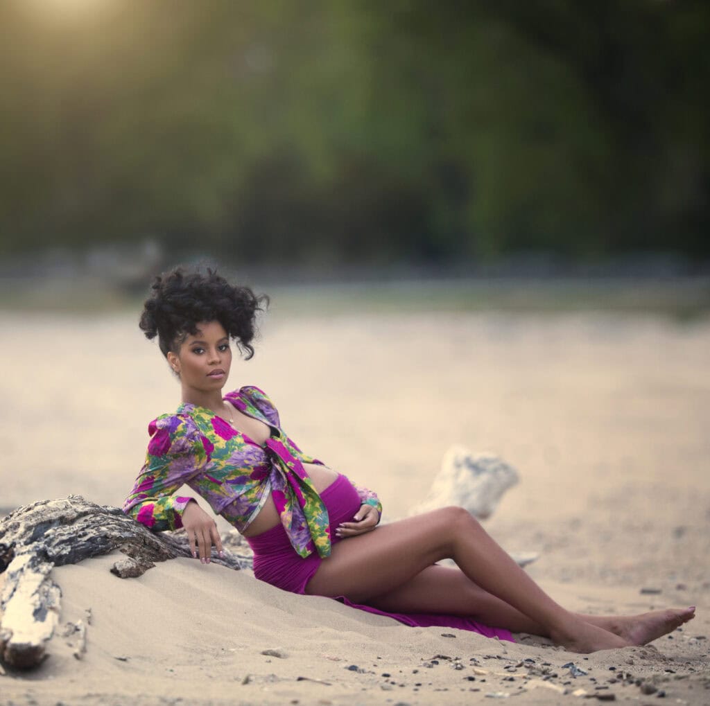 Beautiful African American woman wearing colorful top and skirt showing her pregnant belly while sitting on the beach resting against a large piece of drift wood. The sun is setting behind her and creates a beautiful golden glow around her. This whimsical photo was taken in Chicago by the best pregnancy photographer Sri from Sri and Jana Photography.