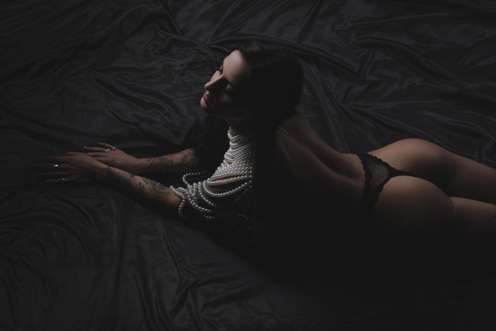 This captivating photograph features a young Italian American woman exuding elegance as she poses in black lingerie atop a bed adorned with lavish black satin sheets. The striking contrast between the lingerie and the sheets creates a visually stunning composition. Additionally, the woman is adorned with exquisite pearls, further adding to the allure of the image. This alluring boudoir photo was taken during a session at a photography studio in the vibrant city of Chicago. The masterful work of the highly skilled boudoir photographer, Sri from Sri and Jana Photography, is evident in the artful composition and attention to detail captured in this photograph.